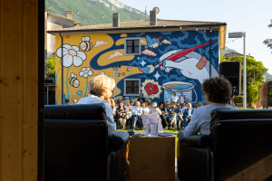 Inaugurazione del murale PACE15×8 al Policaffè di Dolcè: due relatori seduti in primo piano di spalle, pubblico numeroso in giardino e il murale completato di Octoflyart sullo sfondo. L'opera raffigura api, fiori, una colomba e una mano con pennello su sfondo blu e giallo ocra. Sulle colline della Valdadige visibili oltre il tetto.